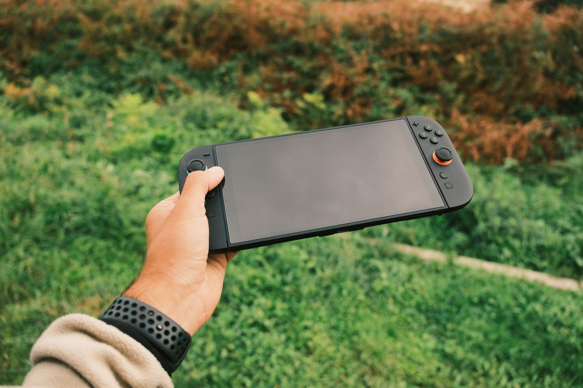 Gros plan de la main d'une personne tenant une console de jeu portable noire sur fond vert extérieur.