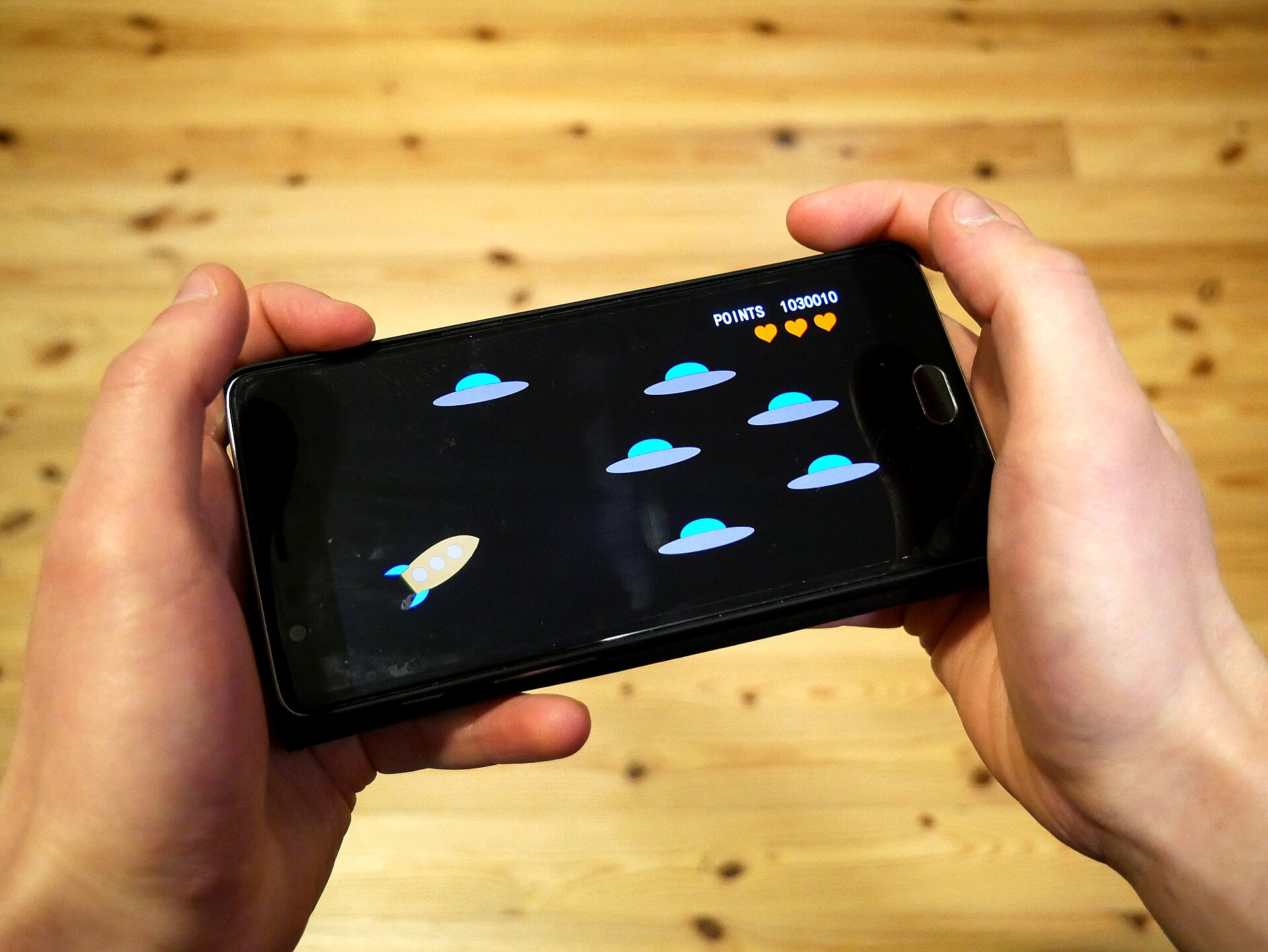 Deux mains tenant un smartphone noir à l'horizontale pour jouer à un jeu de tir spatial, au-dessus d'une table en bois.