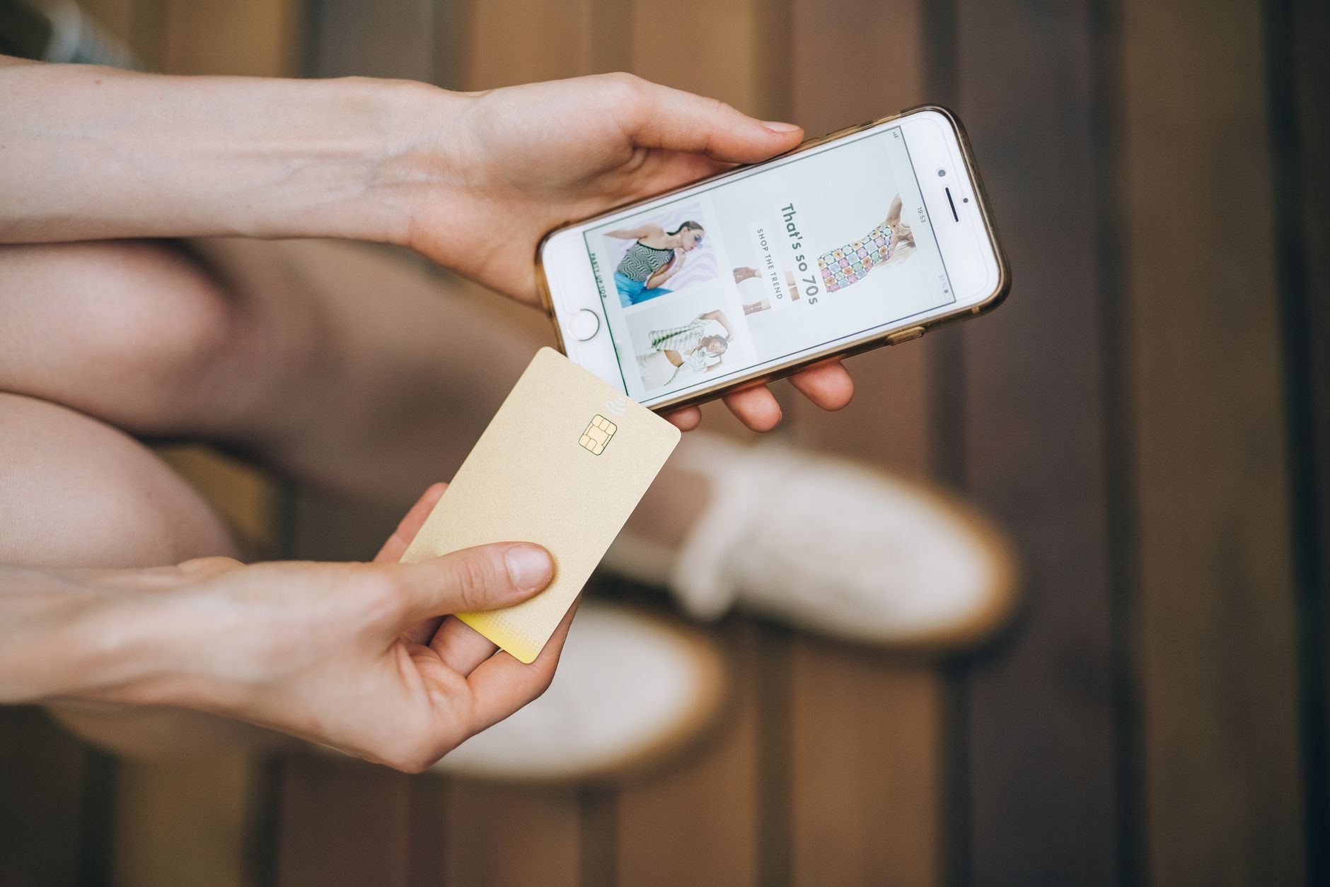Mains tenant un smartphone qui affiche une boutique de mode en ligne et une carte bancaire dorée, au-dessus d’un parquet en bois.