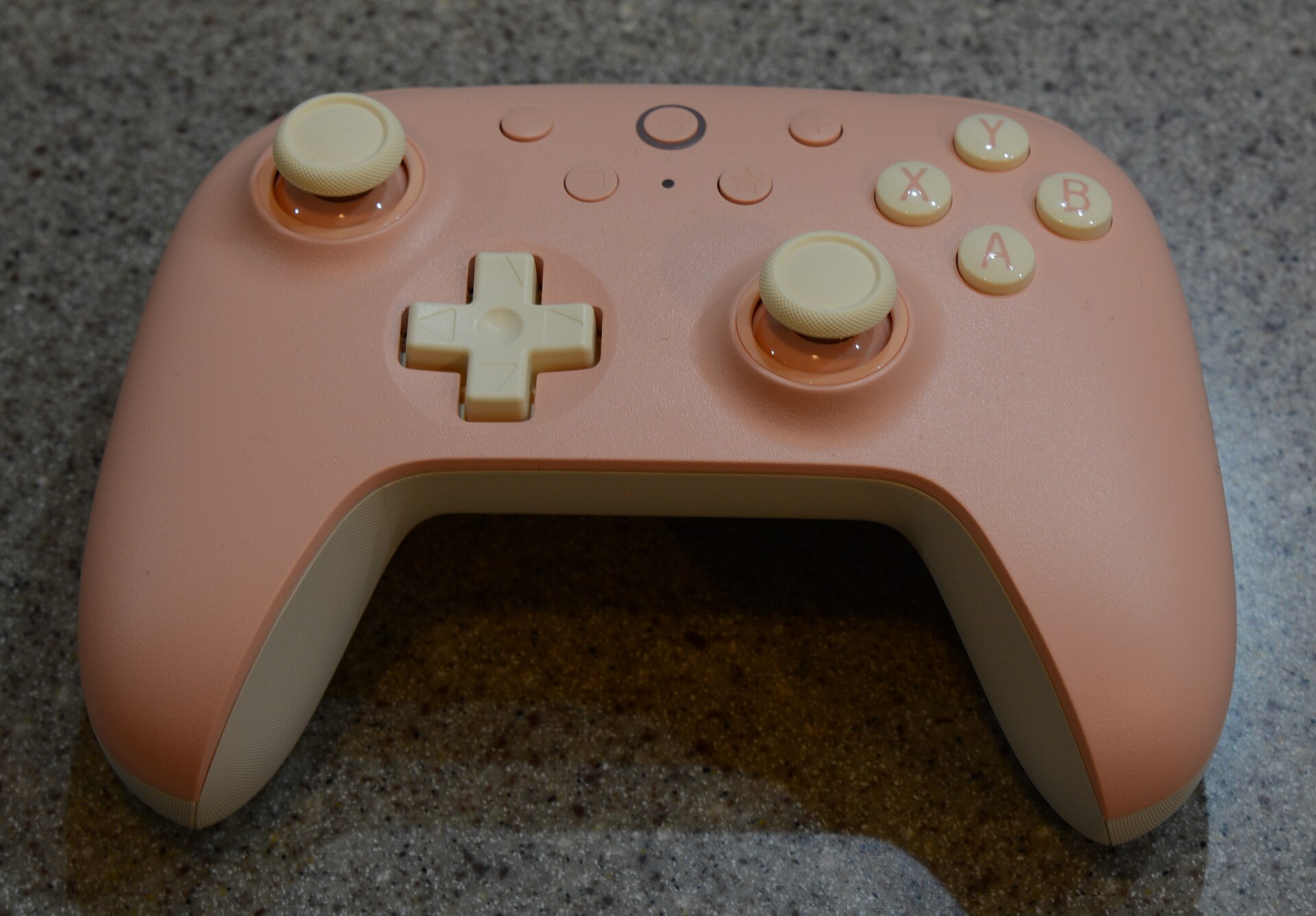 Manette de jeu rose pâle à boutons crème, avec deux joysticks, une croix directionnelle et quatre boutons A, B, X et Y, posée sur un plan de travail granité