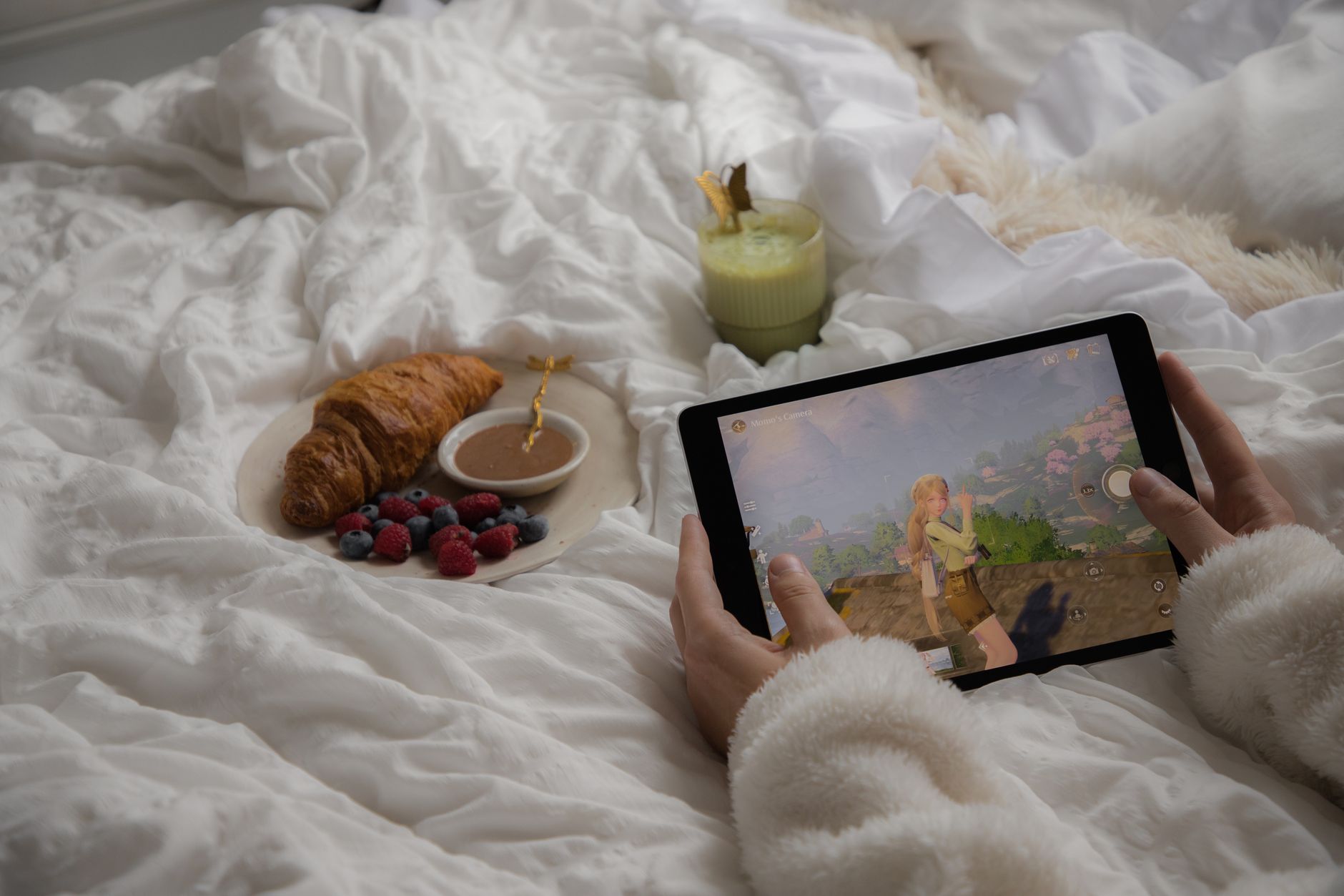 Personne dans un lit tenant une tablette qui affiche un jeu vidéo, avec un croissant, des baies et une boisson verte à côté.