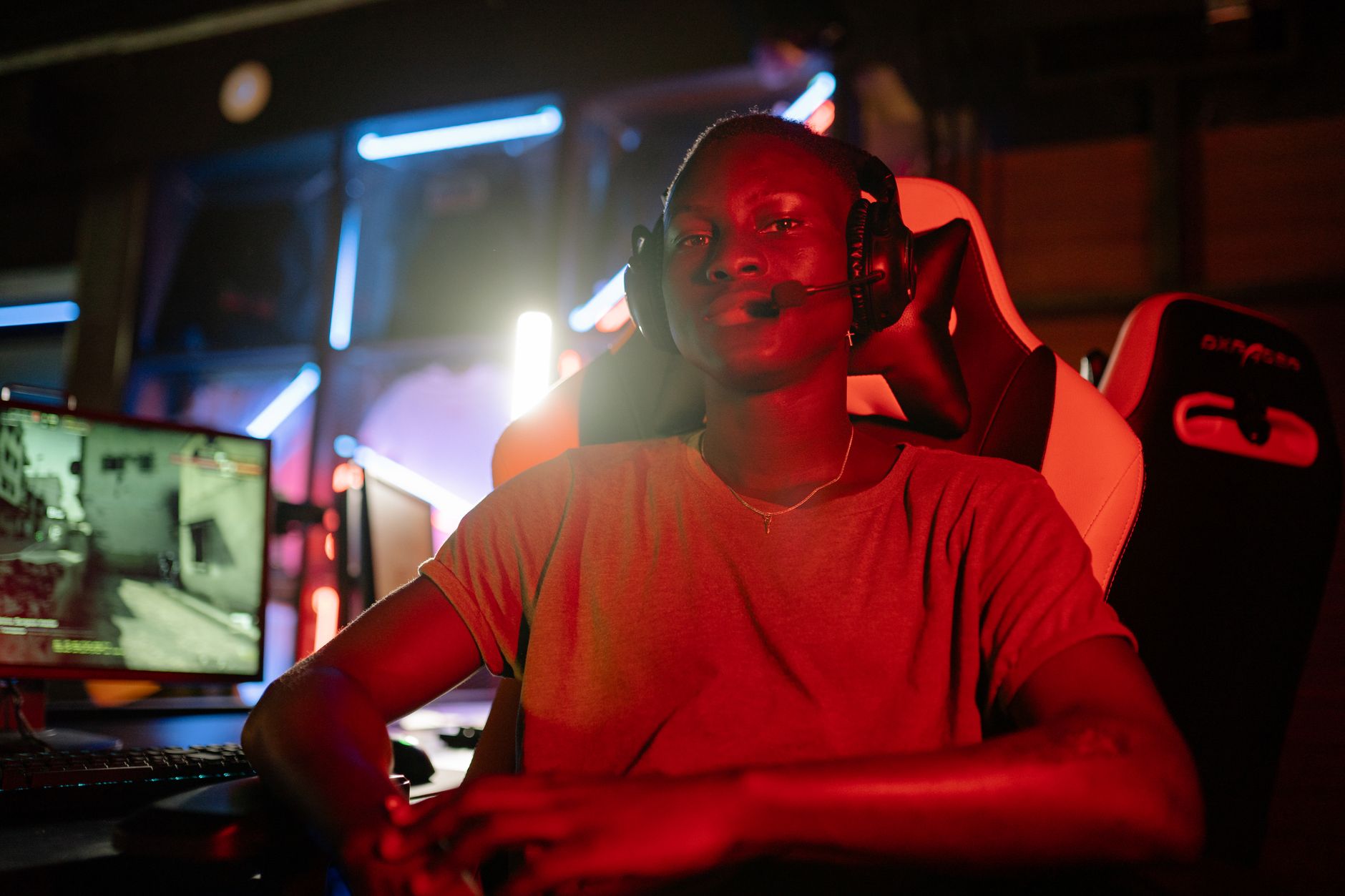 Personne portant un casque-micro noir, assise dans un fauteuil gaming devant un écran et un clavier, dans une pièce sombre éclairée de rouge.